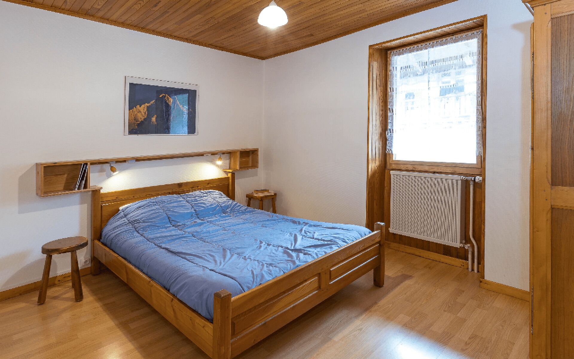 A bedroom with a wooden bed, a nightstand, a small stool, a radiator, and a window with a view of buildings.