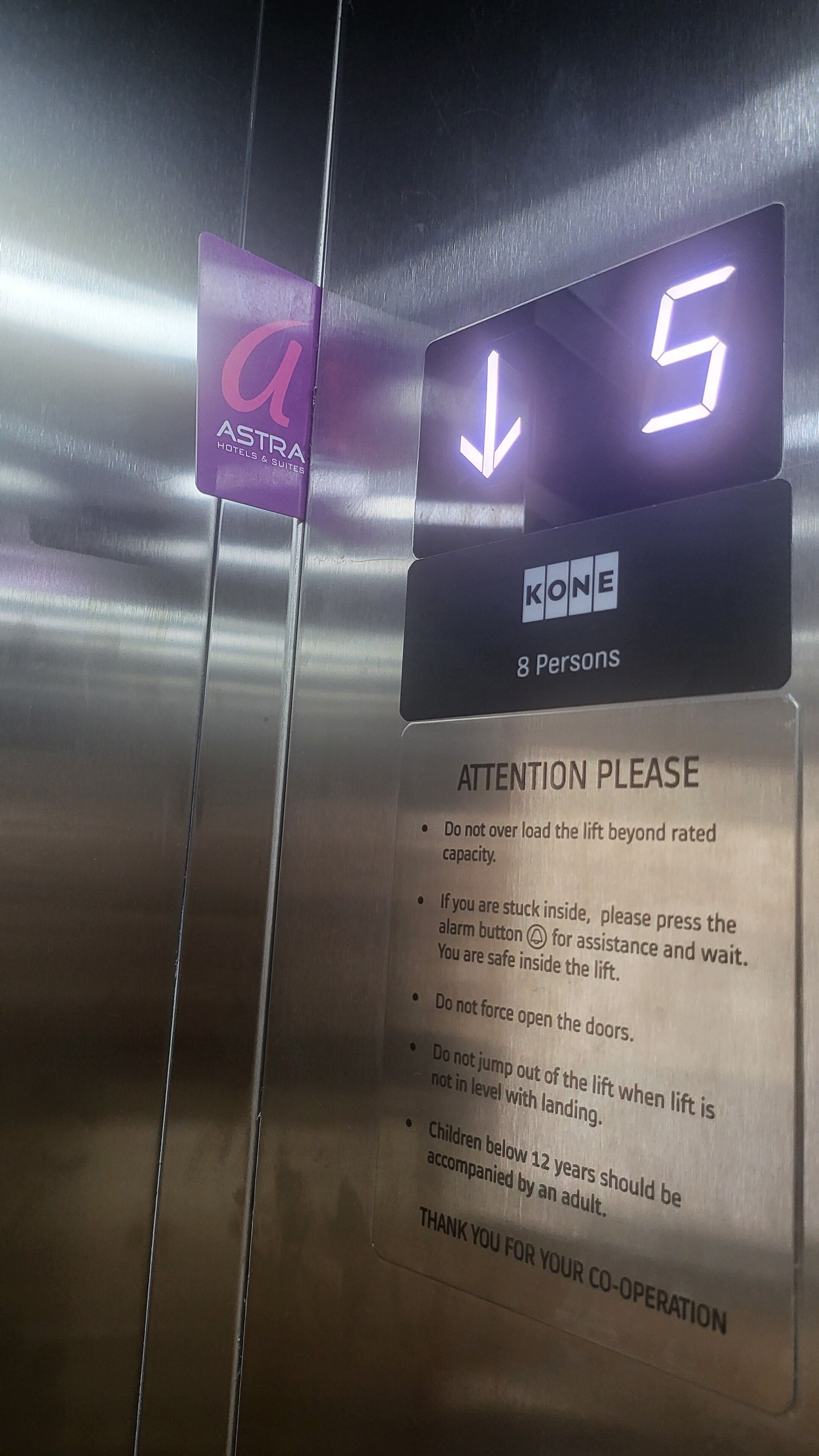 An elevator control panel with a digital display indicating the elevator is on the 5th floor, a purple sign for Astra Hotels & Suites, and safety instructions.