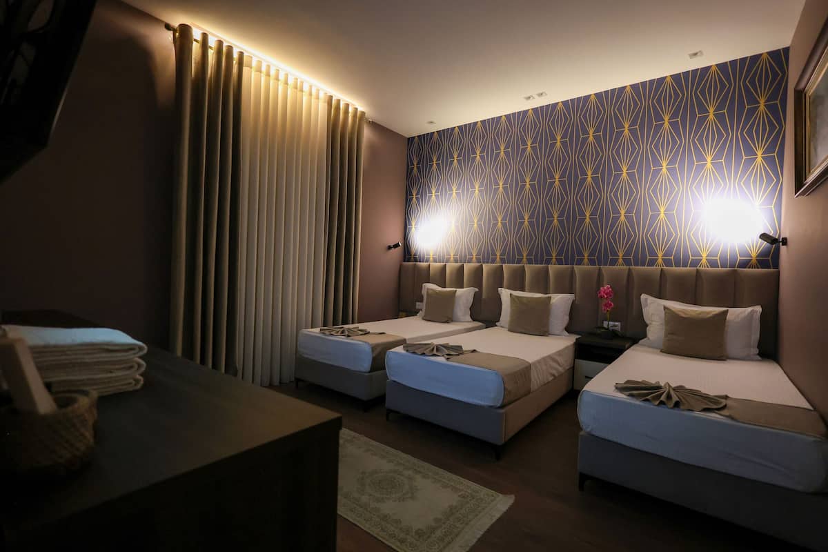Quarto de hotel com duas camas de solteiro, uma janela grande com cortinas, uma mesinha de cabeceira e uma escrivaninha com abajur.