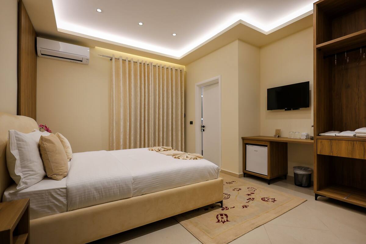 Quarto moderno com cama, mesa de cabeceira, televisão, guarda-roupa e pequena escrivaninha com lixeira.