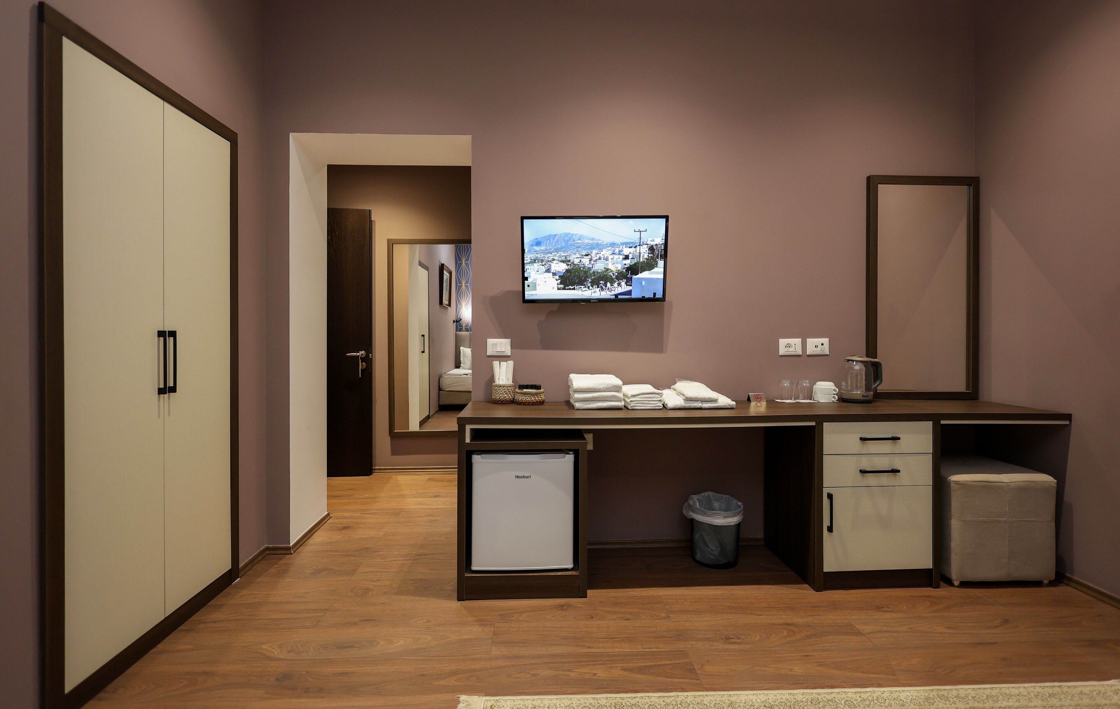 A modern hotel room with a wooden floor, a desk with a flat-screen TV, a small refrigerator, a trash bin, and a mirror.