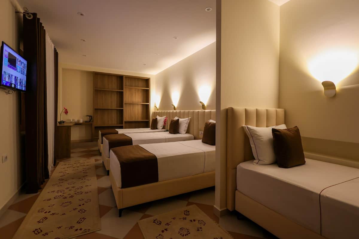 Quarto de hotel com três camas, televisão, armário de madeira e luminárias na parede.