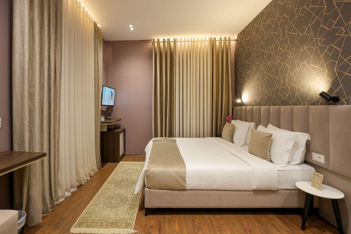 Quarto de hotel com cama, escrivaninha, cadeira, mesinha e televisão na parede.