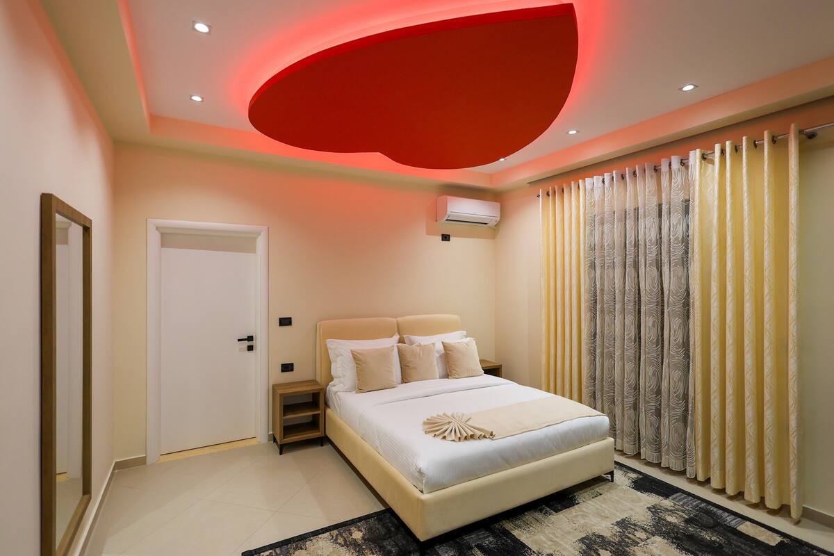 Quarto moderno com uma cama grande, uma mesinha de cabeceira, uma porta e uma janela com cortinas.
