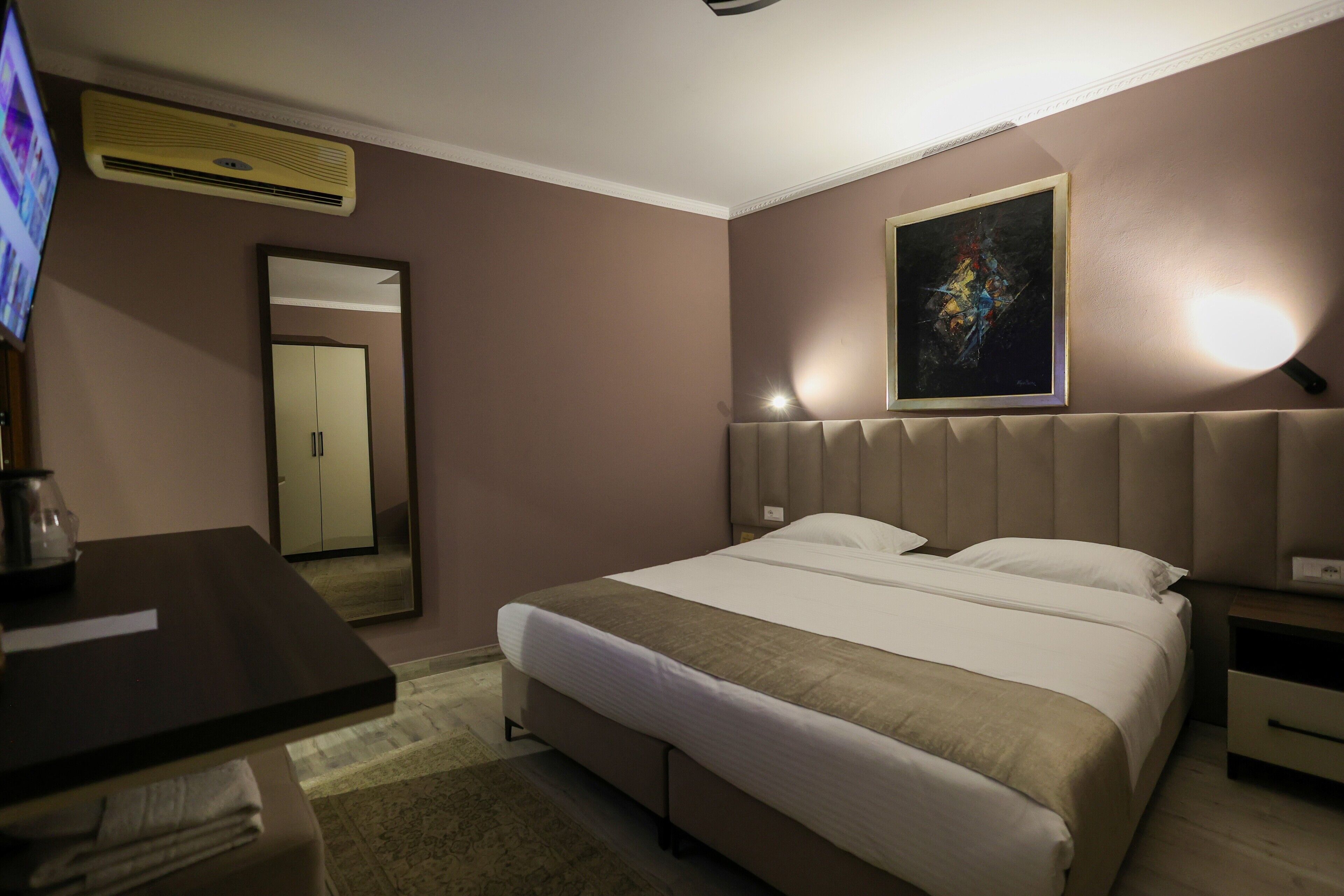 Habitación de hotel con cama, escritorio, silla, televisor, espejo y un cuadro en la pared.