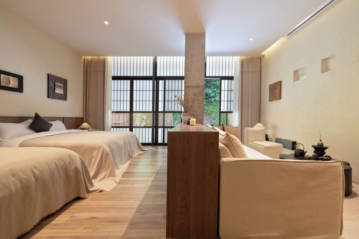 A hotel room with two beds, a wooden table, a sofa, and a view of greenery through large windows.
