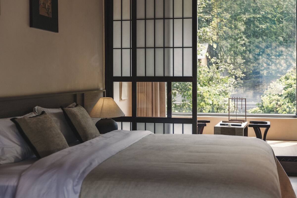 A hotel room with a large bed, a bedside table with a lamp, a view of greenery outside, and a sliding door with a traditional design.