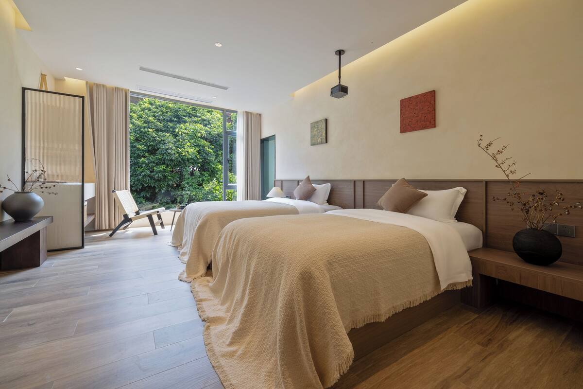 A modern hotel room with two beds, a sitting area, and a view of greenery outside.