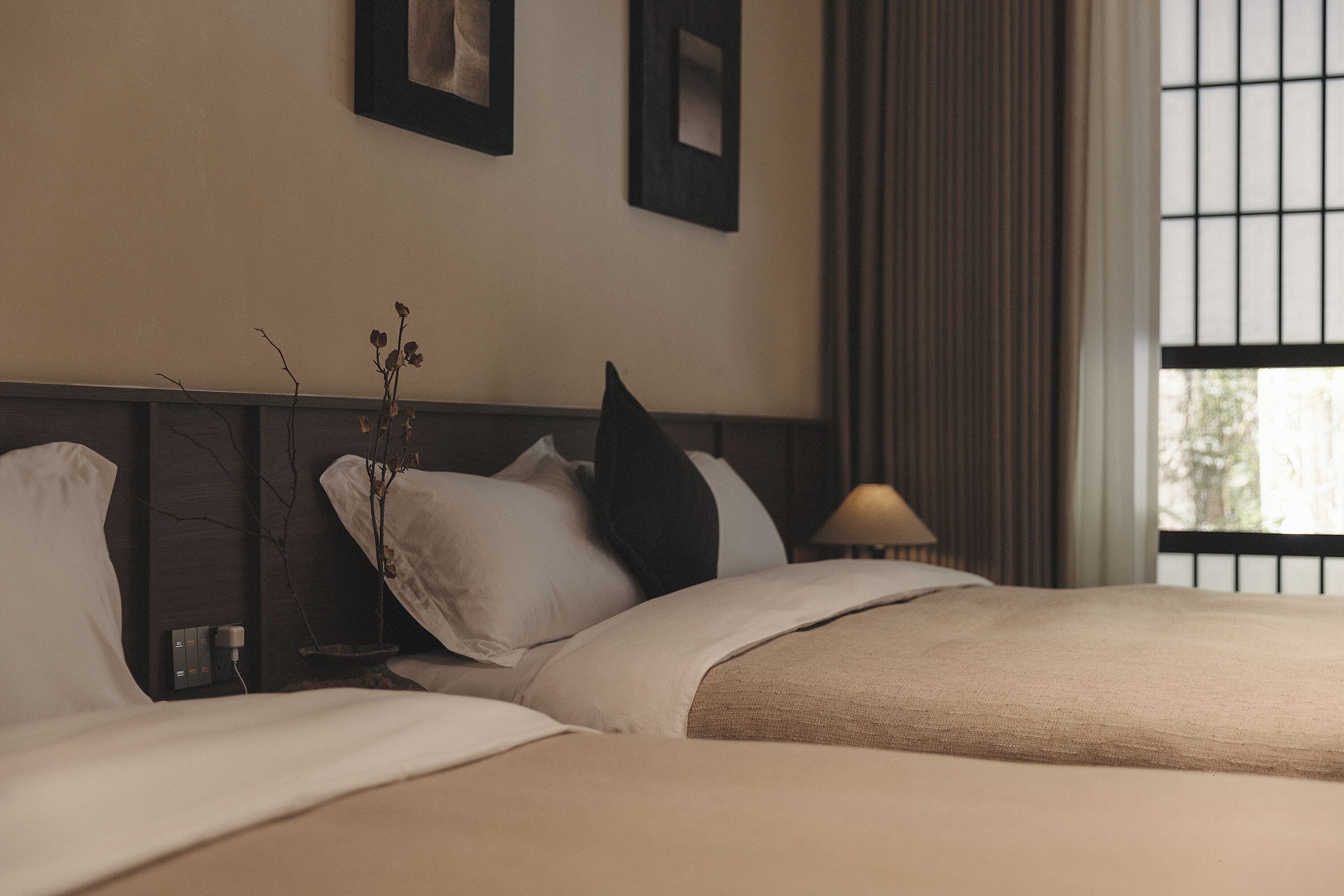 A hotel room with two beds, a bedside table, a lamp, and a window with curtains.