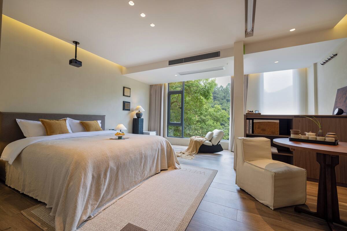 A modern bedroom with a large bed, a bedside table, a chair, and a view of greenery outside.