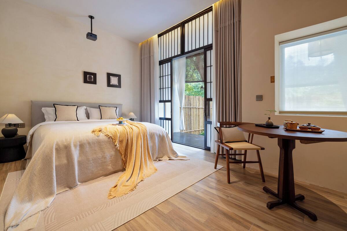 A bedroom with a large bed, a bedside table with a lamp, a chair, a window with curtains, and a view of the outdoors.