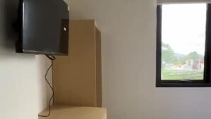 A room with a flat-screen TV mounted on the wall, a wooden bench, and a window with a view of the outside. - Slara Hostel Syariah 1 (Bandar Lampung)