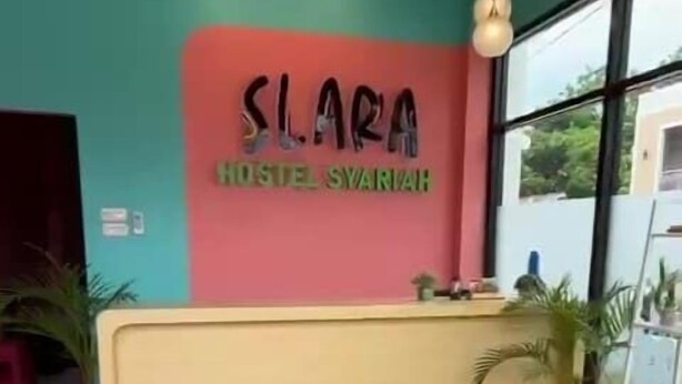 A reception desk with a sign reading "SLARA HOSTEL SYARIVAH" in a room with teal walls and large windows. - Slara Hostel Syariah 1 (Bandar Lampung)