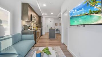 A modern living room with a sofa, a coffee table with a plant, and a kitchen area with cabinets and a TV displaying a beach scene.
