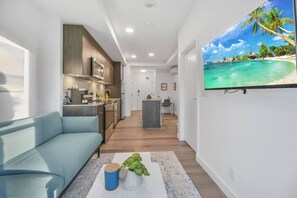 Ein modernes Wohnzimmer mit einer Couch, einem Couchtisch mit einer Pflanze und einer Küchenzeile mit Schränken sowie einem Fernseher, der eine Strandansicht zeigt.