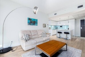 Uma sala de estar moderna com um sofá, uma mesa de centro e uma cozinha ao fundo.