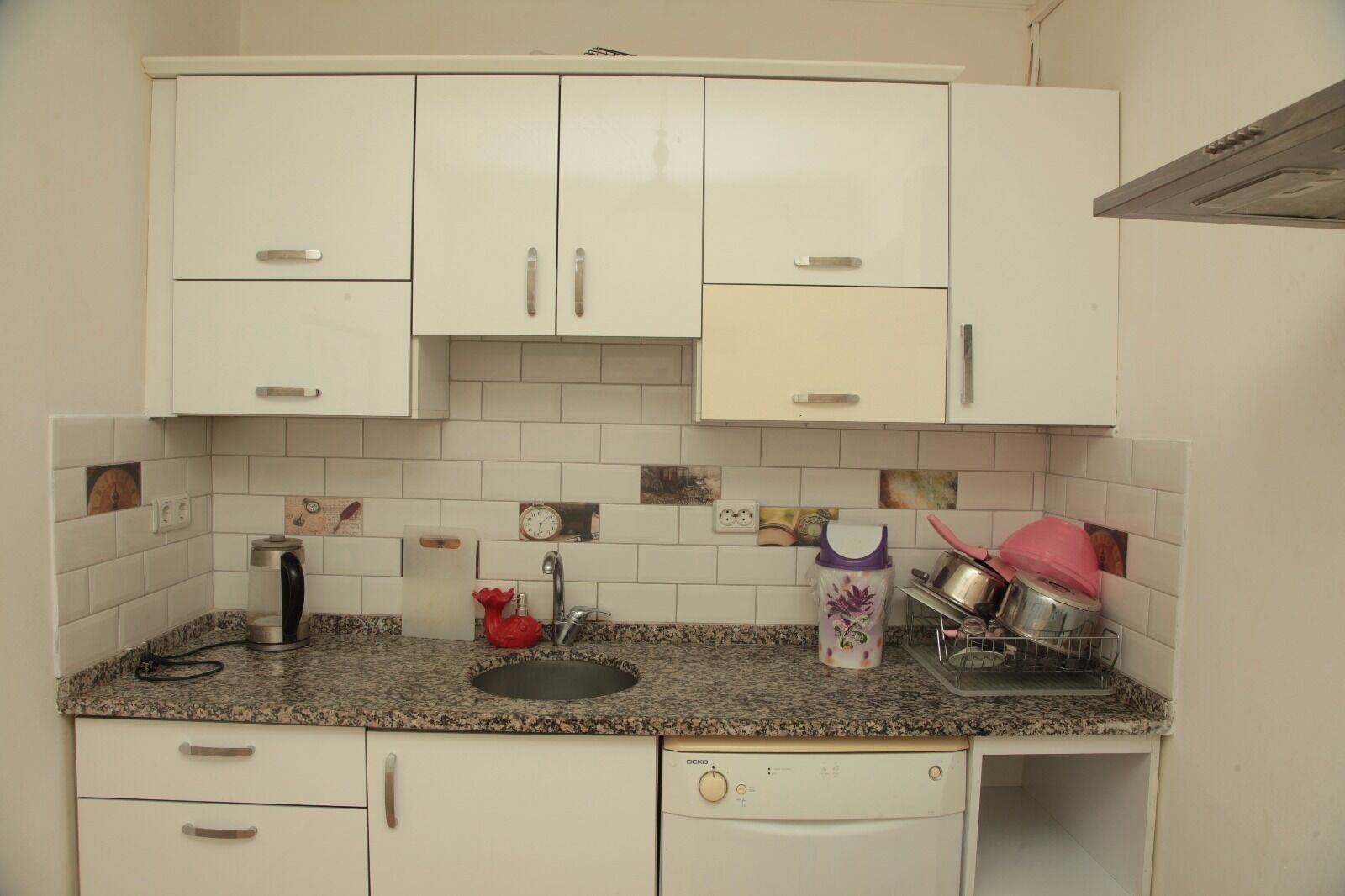 Una cocina compacta con armarios blancos, encimera de granito, un lavabo, un hervidor, un escurreplatos y una lavadora.