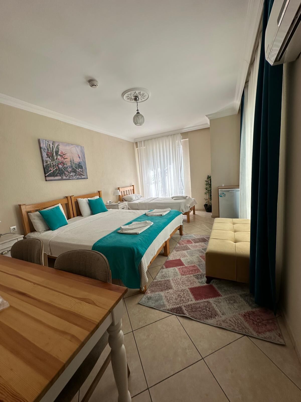 A hotel room with two beds, a wooden desk, a small table, a sofa, a radiator, and a window with curtains.