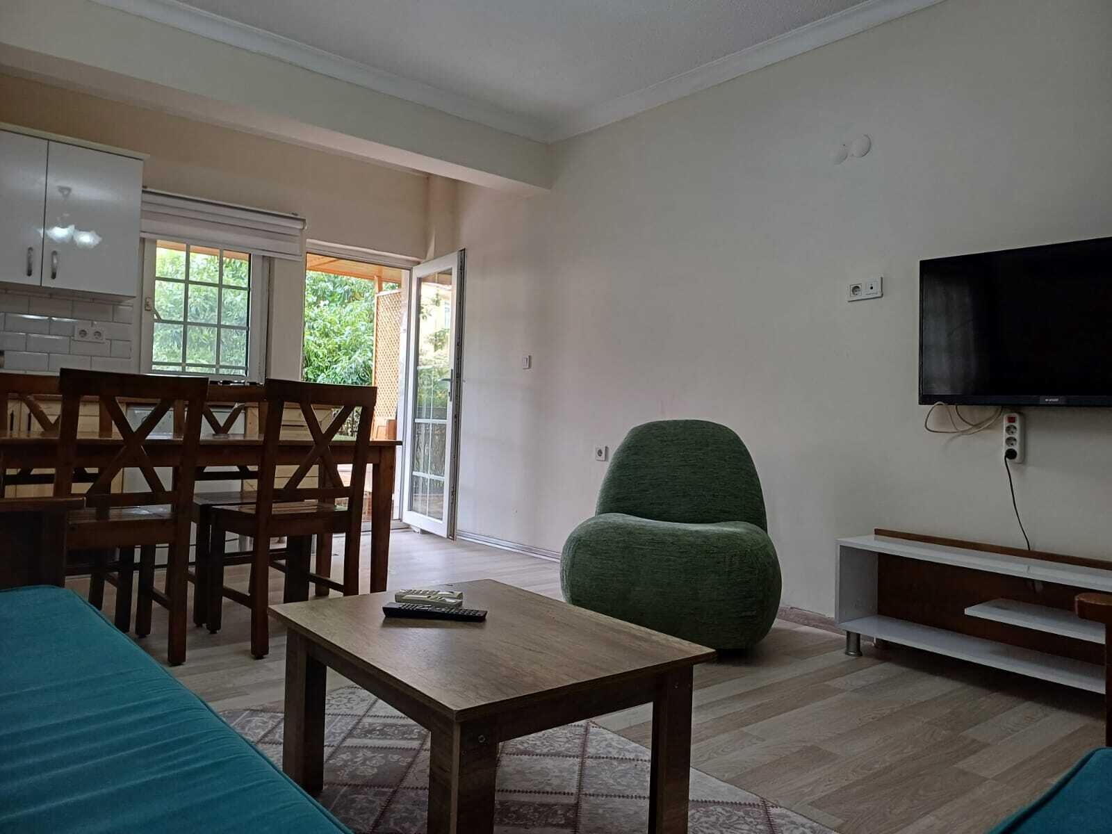Una sala de estar con una mesa de madera, un sillón verde, un televisor montado en la pared y un ventanal corredero que da a un balcón.