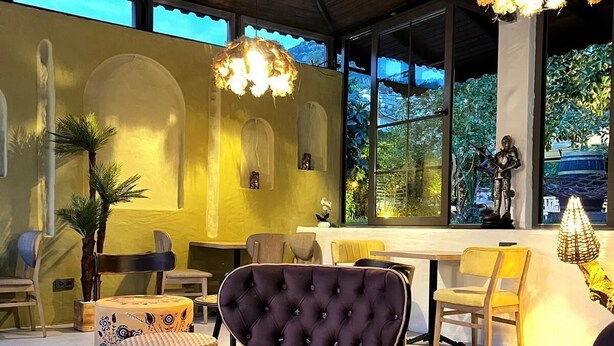 A cozy outdoor seating area with cushioned chairs and tables, a decorative wall, and a large window offering a view of greenery. - Kampüs Garden (Fethiye)