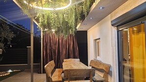 Un patio al aire libre con una mesa y bancos de madera, rodeado de un jardín con plantas colgantes y un accesorio de luz circular.