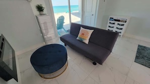 A living room with a sofa, a round ottoman, a TV, a small cabinet, and a view of the beach. - Oceanfront Studio with Sunrise Views & Beach Access (Melbourne Beach)