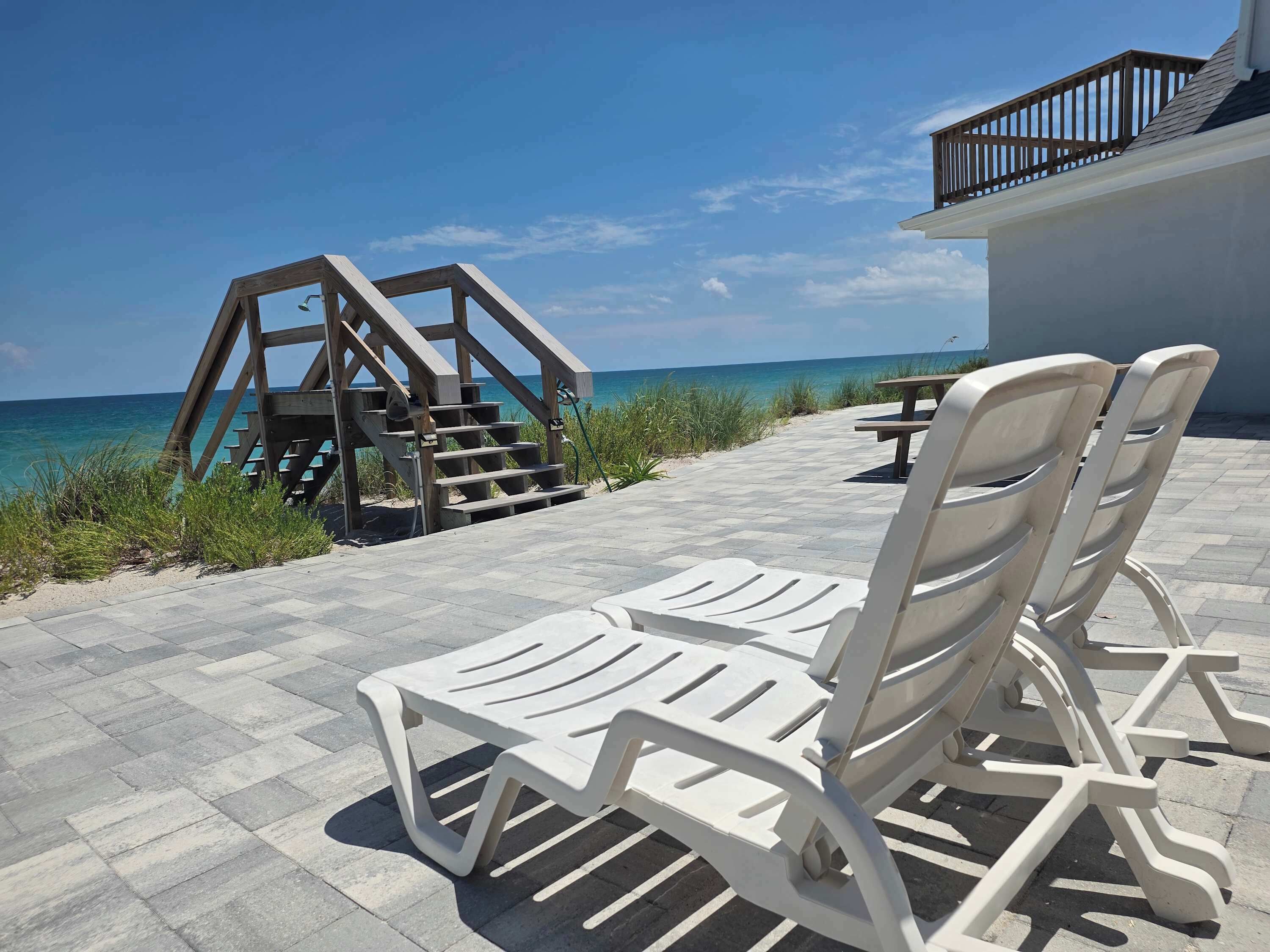 Une terrasse au bord de la plage, avec un escalier en bois menant à une plateforme, des chaises longues en plastique blanc et une vue sur l’océan.