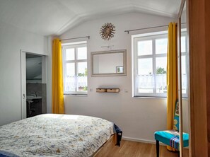 Un dormitori amb un llit, una taula petita, una cadira, una finestra amb cortines grogues, un mirall fixat a la paret i una decoració de sol.