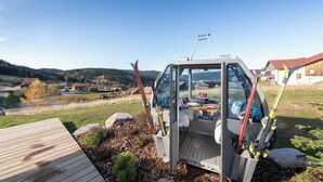 Un véhicule doté d’une paroi latérale transparente et équipé de skis, garé sur une terrasse en bois, d’où l’on peut admirer un quartier résidentiel et des collines en arrière-plan.