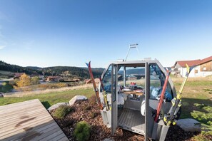 A vehicle with a transparent side, equipped with skis, parked on a wooden deck with a view of a residential area and hills in the background.