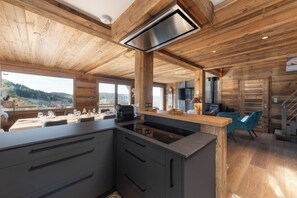 A modern kitchen with a central island, wooden ceiling, and large windows offering a view of the outdoors.