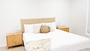 Una cama bien hecha con edredón blanco y manta beige, flanqueada por dos mesitas de noche con lámparas y un pequeño objeto decorativo.