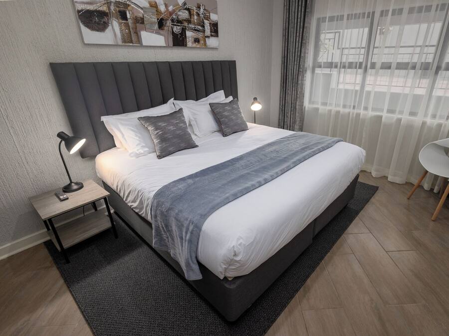 A modern bedroom with a large bed, a bedside table with a lamp, a chair, and a view of the outside through sheer curtains.