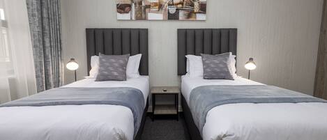 A hotel room with two beds, a nightstand, a wall-mounted artwork, and a window with curtains.