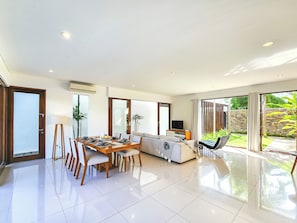 A spacious living area with a dining table, chairs, a sofa, a TV, and a balcony with a view of greenery.
