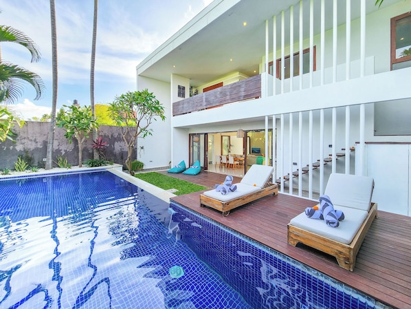 A modern two-story house with a swimming pool, lounge chairs, and a dining area.