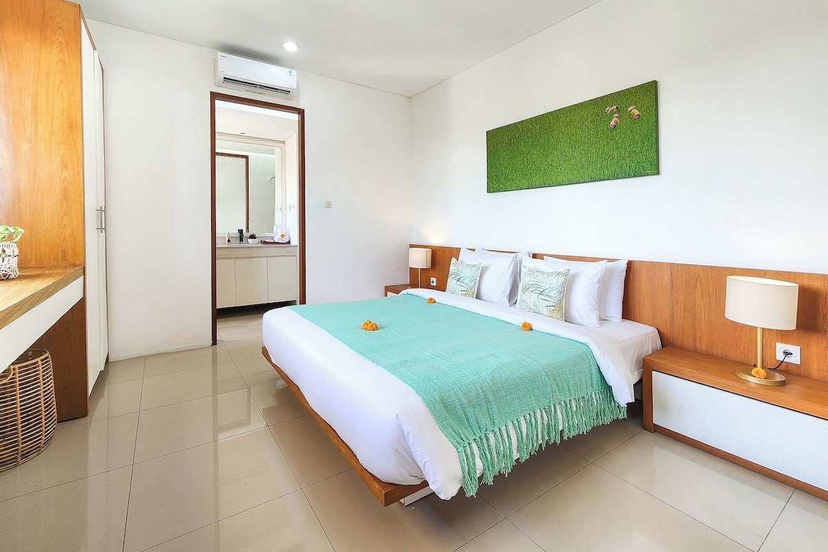 A hotel room with a bed, bedside tables, a mirror, an air conditioning unit, and a green wall decoration.