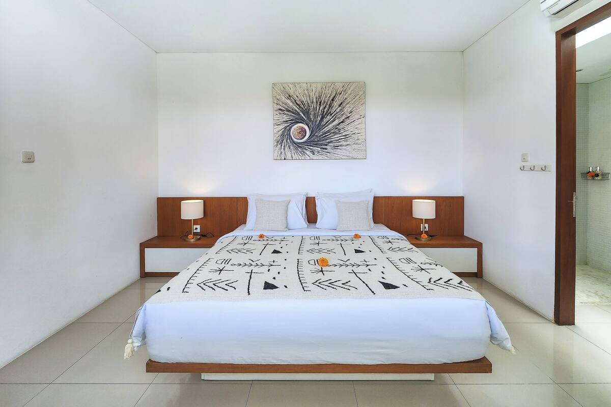 A modern hotel room with a large bed, two bedside tables, a wall-mounted artwork, and a door leading to a bathroom.