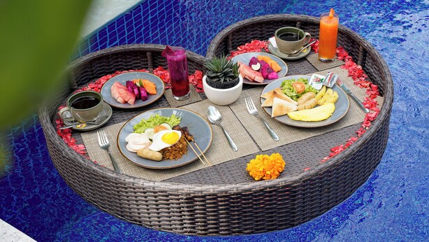 A round wicker table with a variety of dishes, including a plate with eggs, a bowl of soup, and a glass of juice, set on a blue tiled floor.