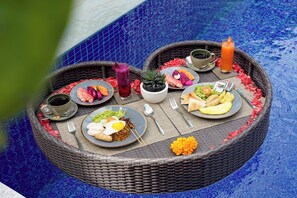 A round wicker table with a variety of dishes, including a plate with eggs, a bowl of soup, and a glass of juice, set on a blue tiled floor. - Villa Abigail Seminyak by Nagisa Bali (Kerobokan)