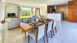 A modern dining area with a long wooden table, cushioned seating, and a kitchen in the background.