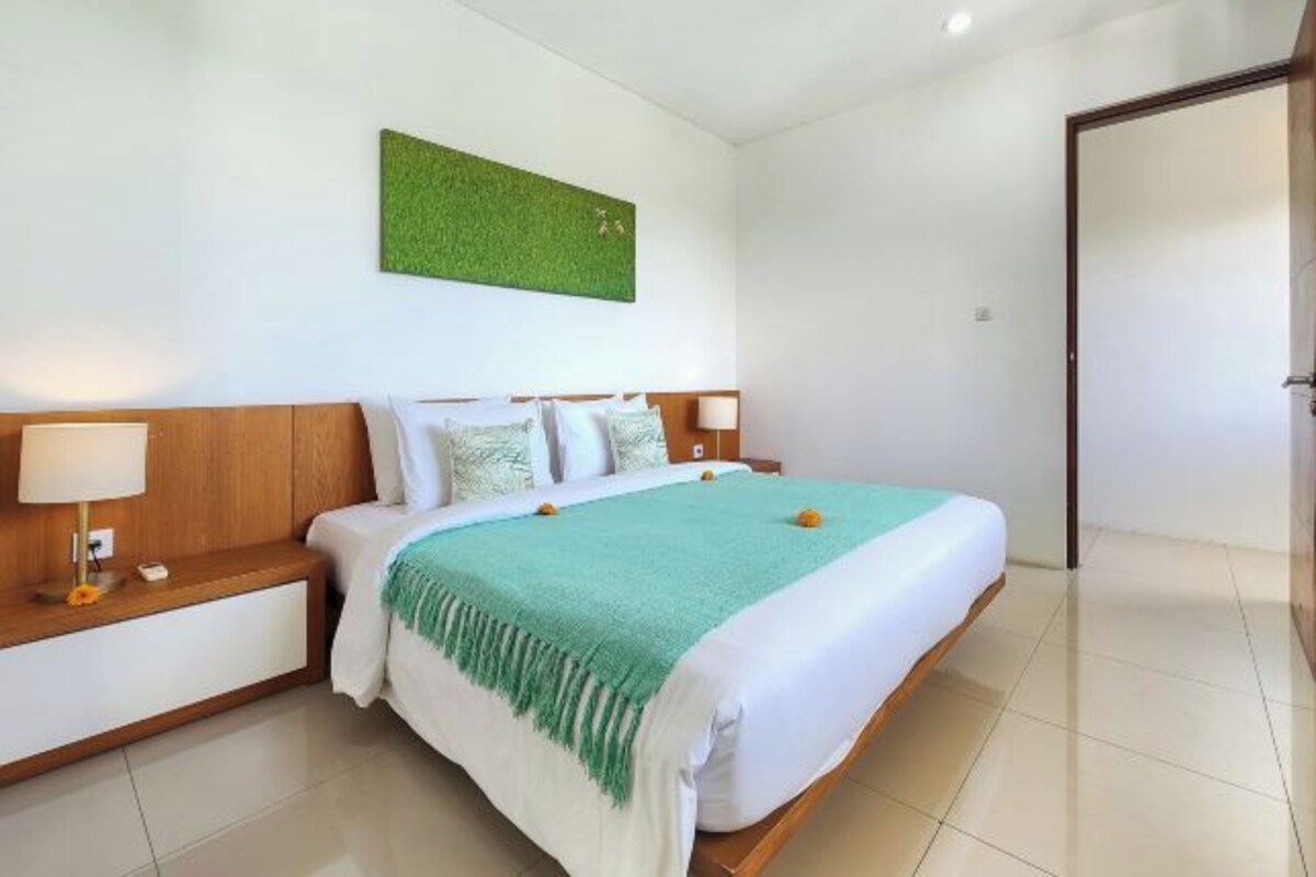 A hotel room with a large bed, a nightstand with a lamp, a green wall decoration, and a wooden door.