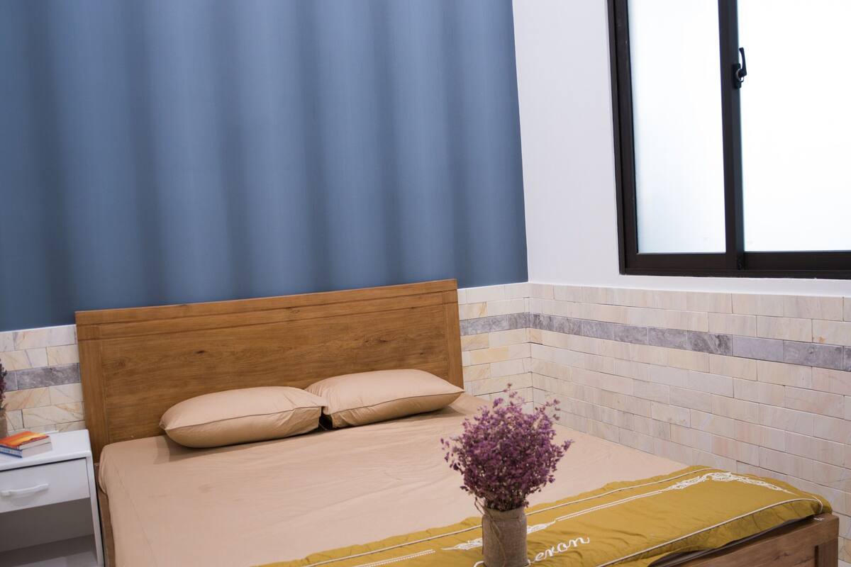 Un dormitorio con cama de madera, una mesita de noche, un florero con flores y una ventana con persianas.