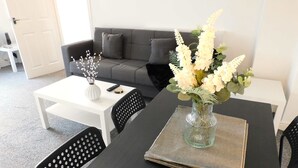 A modern living room with a dark grey sofa, a white coffee table, and a dining table with a vase of flowers. - 4 Bedroom, Newly Refurbished Home in East Kilbride (East Kilbride)