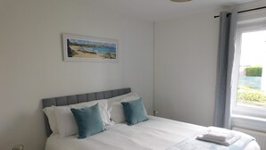 A neatly made bed with white and teal bedding, a small bedside table with a remote control, and a window with curtains. - 4 Bedroom, Newly Refurbished Home in East Kilbride (East Kilbride)