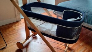 Tumman sinisellä kankaalla päällystetty lastenvaunu, jossa on valkoisia yksityiskohtia, on sijoitettu puulattialle huoneeseen, jonka taustalla näkyy sänky.