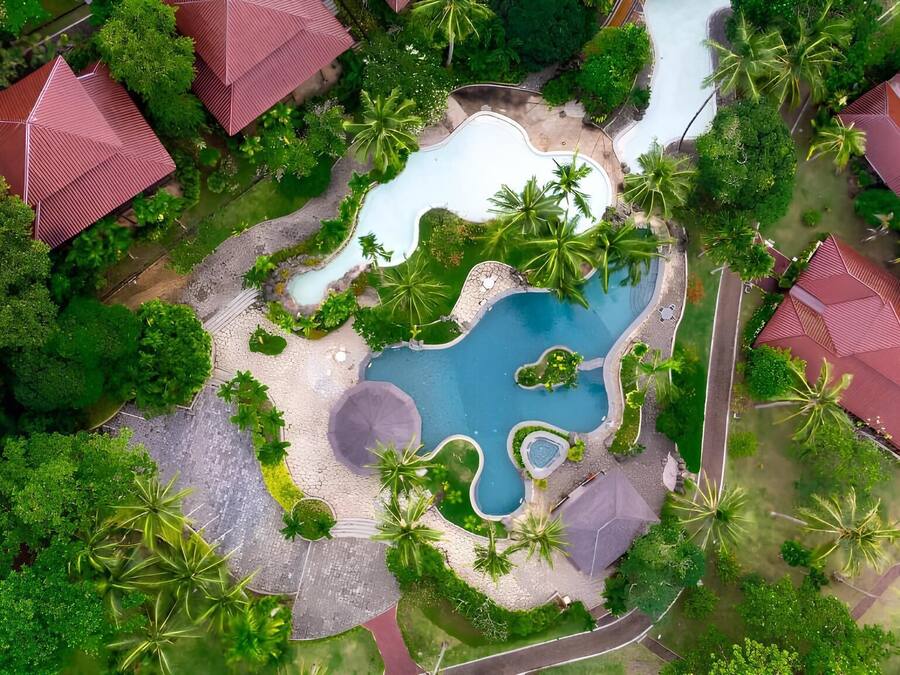 Luchtbeeld van een resort met meerdere gebouwen met rode daken, een centraal gelegen zwembad en omliggend weelderig groen.