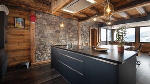 Une cuisine moderne dotée d’un grand îlot central, d’un mur en pierre, d’un plafond en bois et de grandes fenêtres.