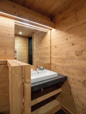 A wooden cabin interior with a bathroom featuring a white sink, a mirror, and a wooden bench.