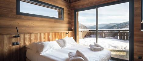 A wooden cabin bedroom with a large bed, a window, and a view of a snowy landscape.